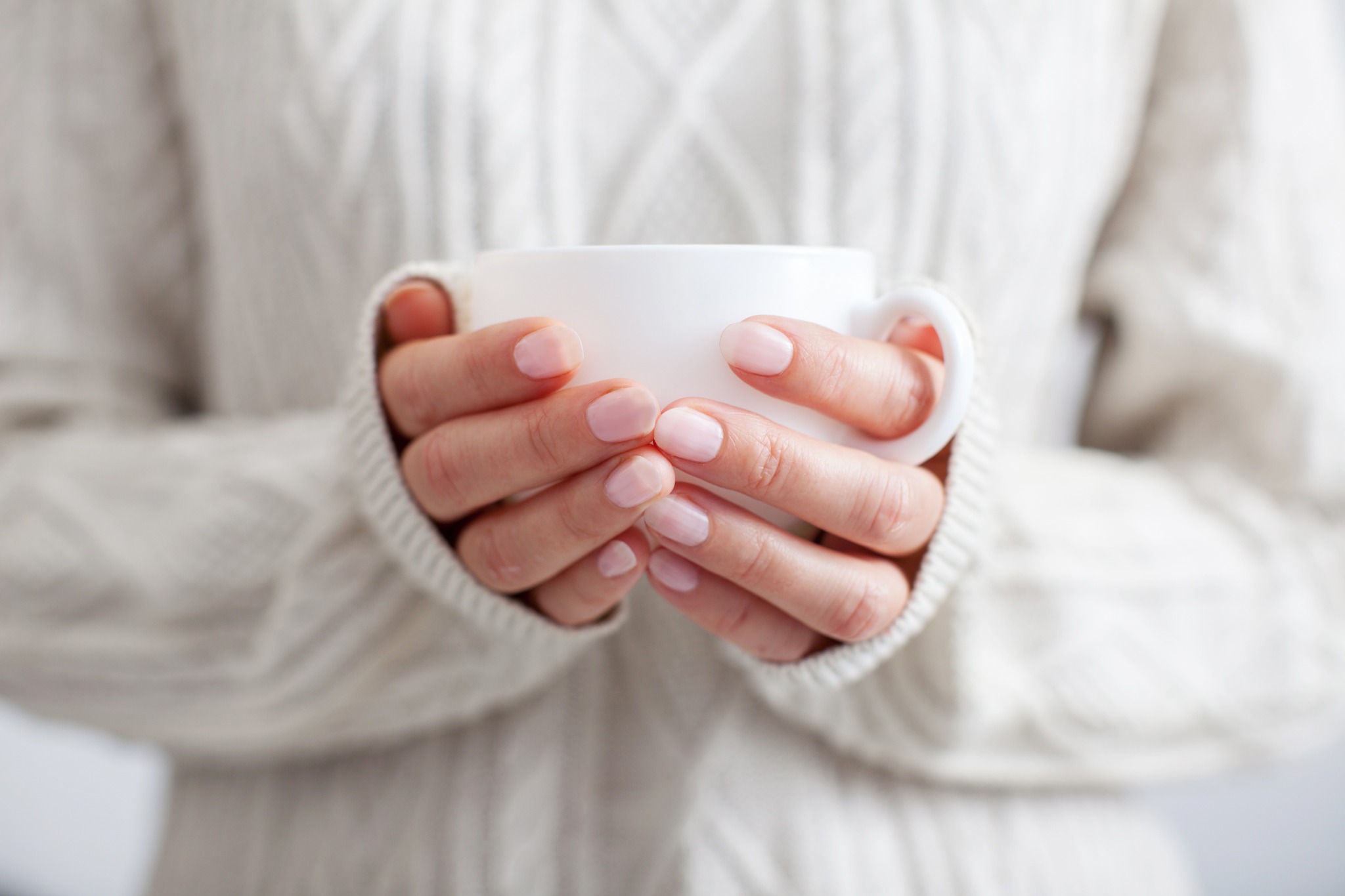 A woman in a sweater holding a white mug to warm her cold hands.