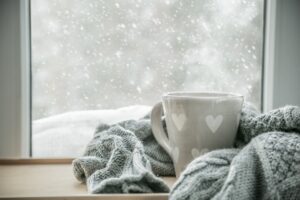 A grey mug with hearts wrapped in a scarf sitting in front of a window with a snowy background.