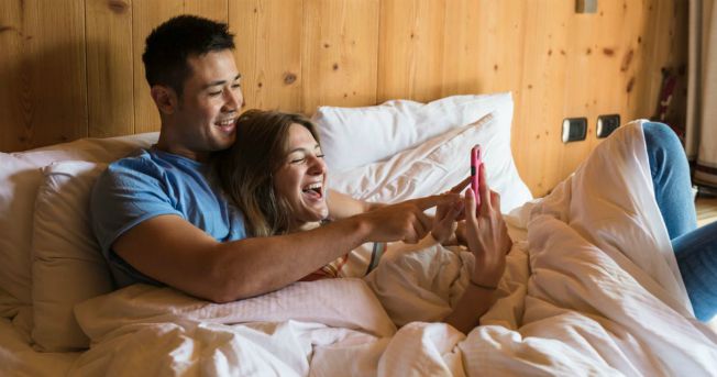 man and woman laying in bed holding a phone and smiling while looking at it