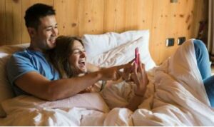 man and woman laying in bed looking at phone screen smiling